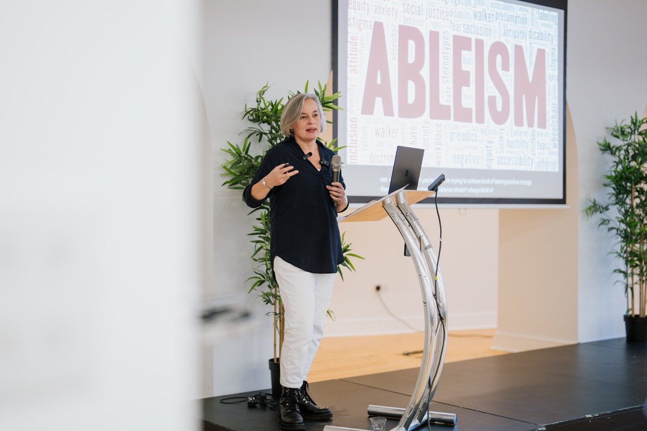 Speaker presenting beside a lectern in front of a screen displaying the word Ableism